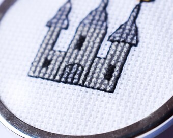 May include: A round, silver-toned pin with a cross-stitched design of a castle. The castle is rendered in black thread on a white background, with three towers topped with small orange flags. The pin has a polished, metallic edge.