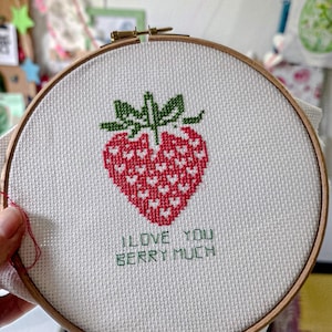Puede incluir: Un bastidor de bordado circular de madera que contiene tela blanca con un diseño de punto de cruz de una fresa roja con pequeños corazones blancos. Debajo de la fresa, el texto I LOVE YOU BERRY MUCH aparece en letras verdes.