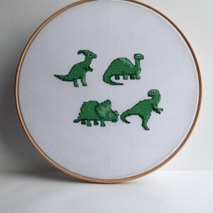 May include: A cross-stitch embroidery hoop featuring four green dinosaur designs on white fabric. The dinosaurs are various species, each with unique shapes and poses. The hoop is made of light-colored wood, creating a simple, handmade aesthetic.
