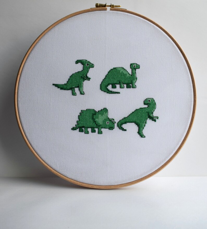 May include: A cross-stitch embroidery hoop featuring four green dinosaur designs on white fabric. The dinosaurs are various species, each with unique shapes and poses. The hoop is made of light-colored wood, creating a simple, handmade aesthetic.