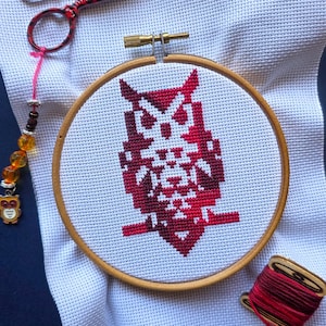 Puede incluir: Un diseño de búho de punto de cruz en un bastidor de bordado de madera. El búho es rojo y blanco sobre tela blanca. Tijeras de bordar rojas y negras, un dije de cuentas rosa y una bobina de hilo rojo también están presentes.
