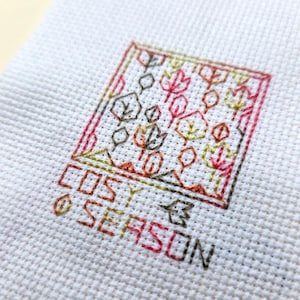 May include: A close-up of a cross-stitch design on white fabric. The design features a square border with various leaf shapes in shades of red, orange, brown, and green. The words "COSY SEASON" are stitched below the square.