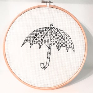 May include: A black and white cross-stitch embroidery of an umbrella with various geometric patterns. The embroidery is set in a round wooden hoop, creating a finished piece of textile art.