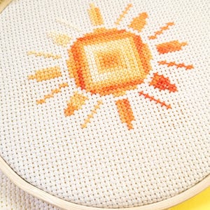 Könnte beinhalten: Ein Kreuzstich-Stickrahmen mit einem Sonnendesign in Orange- und Gelbtönen. Die Sonne ist auf cremefarbenem Stoff gestickt, der in einem Holzrahmen gehalten wird. Ein Strang orange-gelben Garns ist ebenfalls sichtbar.