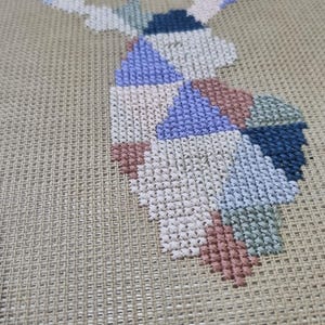 May include: A cross-stitch design of a deer head in various colors, including blue, pink, and green. The needlework is on a tan canvas, showcasing the detailed stitches and geometric patterns. This handmade piece is perfect for home decor.