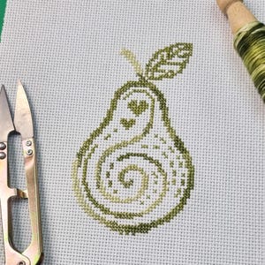 May include: Cross-stitch embroidery of a green pear with a heart design and a spiral pattern. The pear is on white fabric, with scissors and a spool of green thread in the background.