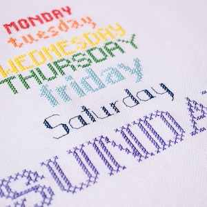May include: A white fabric with cross-stitched text in various colors. The days of the week are displayed, with "MONDAY" in red, "tuesday" in orange, "WEDNESDAY" in yellow, "THURSDAY" in green, "FRIDAY" in light blue, "Saturday" in dark blue, and "SUNDAY" in purple.