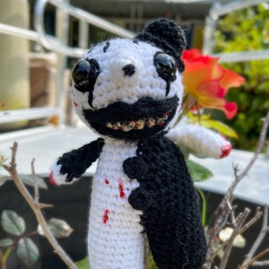 May include: A crocheted plush toy with a white body and black arms and legs. The toy has a black and white face with a wide, toothy grin and red paint splatters on its body.