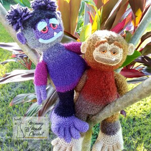 May include: Two crocheted plush toys, a purple and black monster and a brown and red monkey, are sitting on a tree branch. The monster has a purple and pink body, black legs, and a purple and gray head with a purple and white pom-pom on top. The monkey has a brown head, a red and brown body, and brown legs. The toys are both sitting on a tree branch with green grass and red leaves in the background. The image includes the text "Mommy Made Crochet".