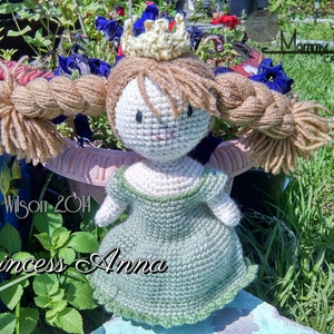 May include: A crocheted doll dressed as Princess Anna from the movie Frozen. The doll has a green dress, a gold crown, and long brown braids. The doll is sitting on a blue and white surface with green grass in the background.