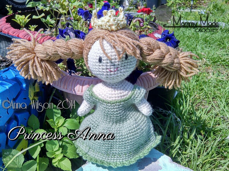May include: A crocheted doll dressed as Princess Anna from the movie Frozen. The doll has a green dress, a gold crown, and long brown braids. The doll is sitting on a blue and white surface with green grass in the background.