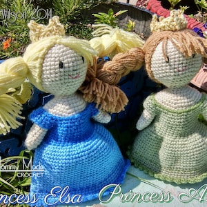 May include: Two crocheted dolls dressed as princesses. The doll on the left is wearing a blue dress and a gold crown. The doll on the right is wearing a green dress and a gold crown. Both dolls have blonde hair and are sitting on a wooden surface. The text "Princess Elsa Princess Anna" is written below the dolls.