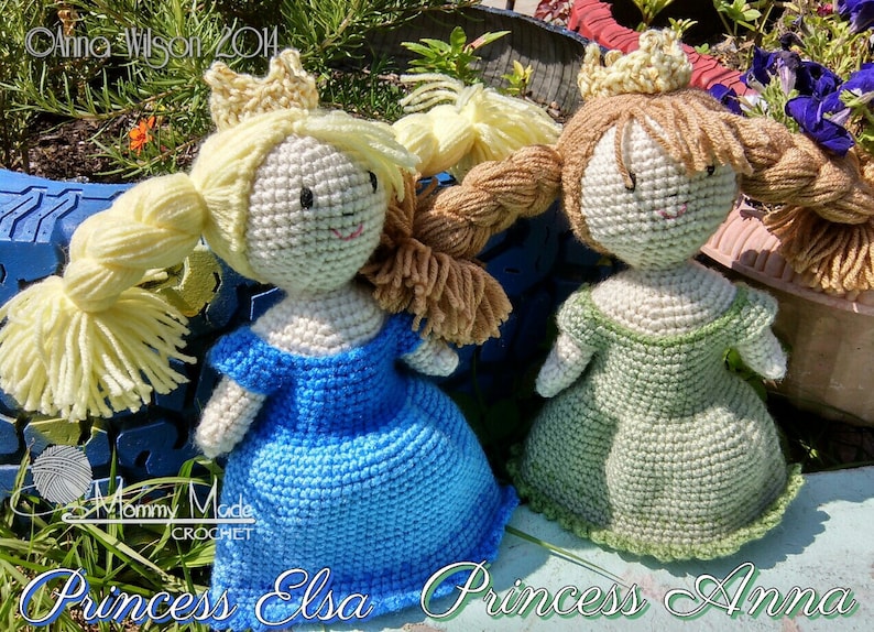 May include: Two crocheted dolls dressed as princesses. The doll on the left is wearing a blue dress and a gold crown. The doll on the right is wearing a green dress and a gold crown. Both dolls have blonde hair and are sitting on a wooden surface. The text "Princess Elsa Princess Anna" is written below the dolls.