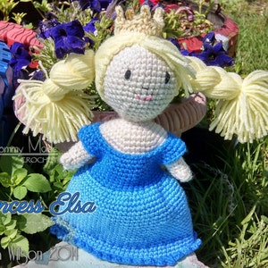 May include: Crocheted doll of Princess Elsa from the movie Frozen, wearing a blue dress and a gold crown. The doll has blonde hair and a smiling face.
