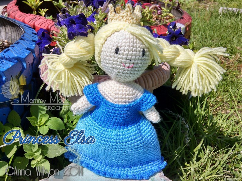 May include: Crocheted doll of Princess Elsa from the movie Frozen, wearing a blue dress and a gold crown. The doll has blonde hair and a smiling face.