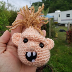 Può includere: Un giocattolo di peluche lavorato a uncinetto a forma di testa di bambino con capelli in lana marrone, pelle rosa, occhi neri e un sorriso con i denti.