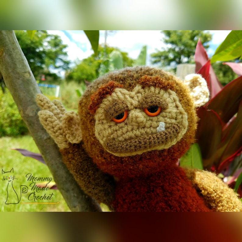 May include: A crocheted monkey toy with brown fur, orange eyes, and a white nose. The monkey is holding onto a branch with one hand and is looking at the camera.