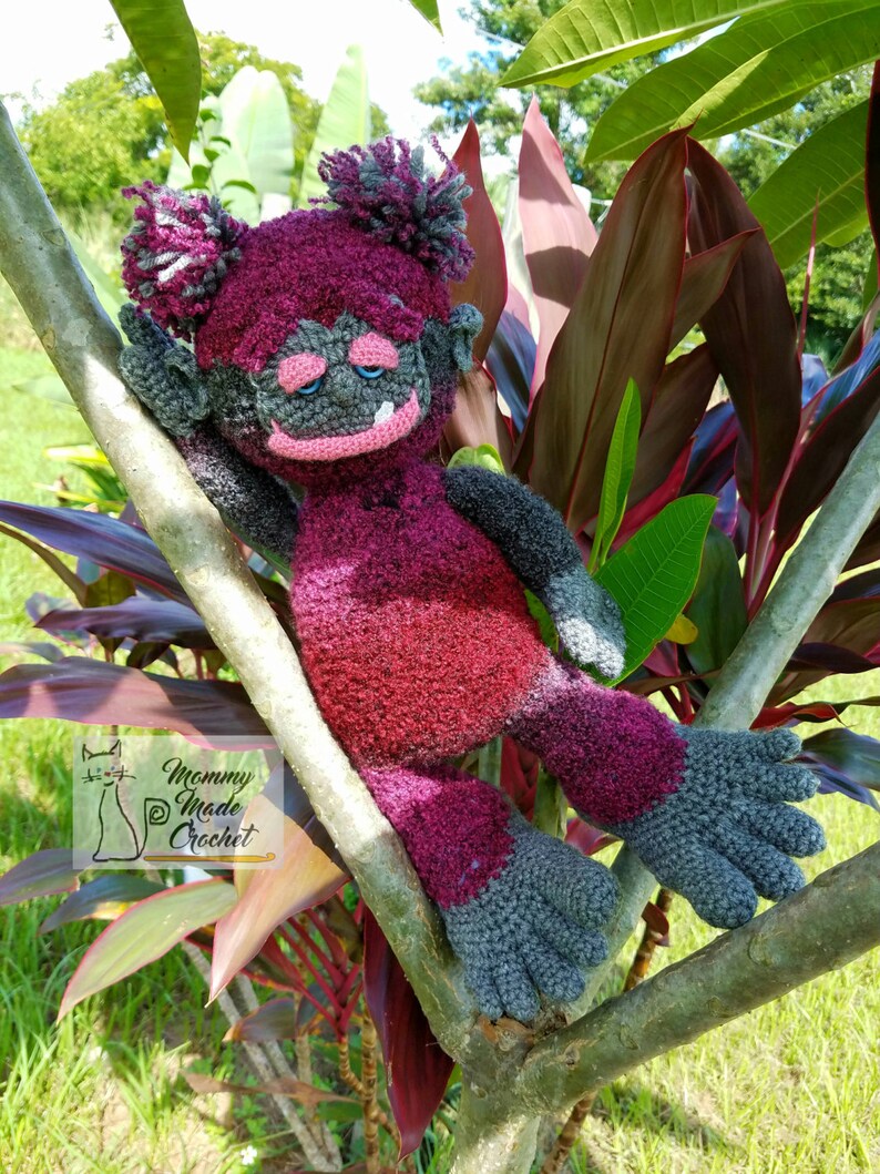 May include: A crocheted purple and gray troll doll with a sad expression, sitting on a branch of a tree with red leaves. The doll has a long, purple body and gray arms and legs. The doll's hair is a fluffy purple and gray pom-pom. The doll's eyes are closed and its mouth is slightly open. The doll's nose is a small, gray bump. The doll's ears are small and gray. The doll's fingers and toes are all separate and have a gray color. The doll is sitting on a branch of a tree with red leaves. The tree is in a garden setting. The doll is made by "Mommy Made Crochet".
