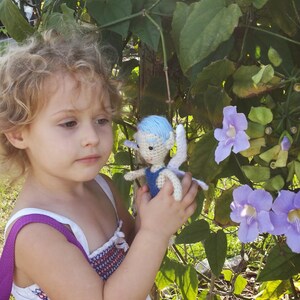 May include: A young girl with curly blonde hair is holding a crocheted fairy doll with blue wings and a light blue dress. The fairy doll is holding a small flower. The girl is wearing a white dress with a purple strap and a purple backpack. The background is a green leafy vine with purple flowers.