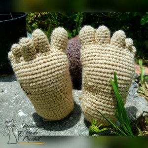 May include: A pair of crocheted beige foot warmers with five toes each. The foot warmers are lying on a gray surface with green grass in the background.