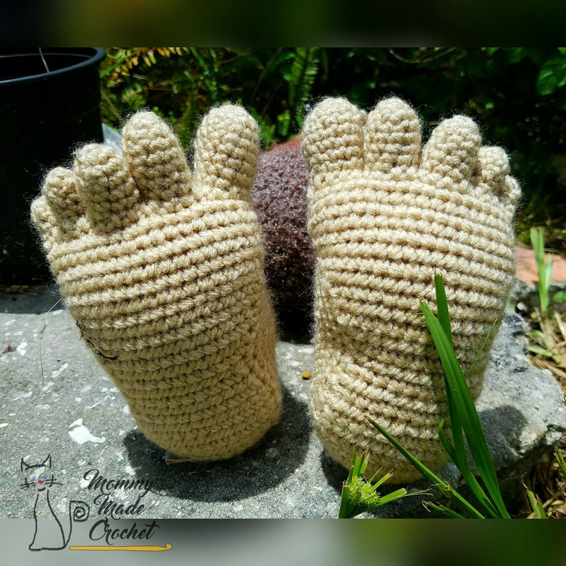 May include: A pair of crocheted beige foot warmers with five toes each. The foot warmers are lying on a gray surface with green grass in the background.