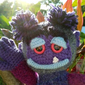 May include: A purple crocheted plush toy with blue eyes and a blue mouth. The toy has two purple yarn buns on top of its head. The toy is holding its arm up in the air.