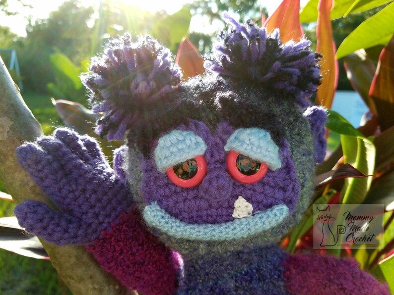 May include: A purple crocheted plush toy with blue eyes and a blue mouth. The toy has two purple yarn buns on top of its head. The toy is holding its arm up in the air.