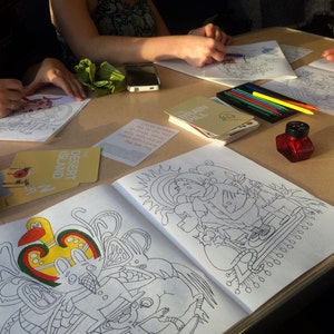 May include: A table with several coloring books and coloring supplies. The coloring books have black and white illustrations of people, animals, and objects. The coloring supplies include colored pencils, markers, and a bottle of ink.