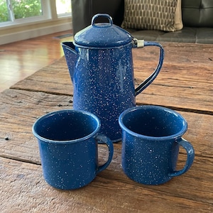 May include: A blue speckled enamelware coffee pot with a lid and two matching mugs. The set is on a rustic wooden table. The coffee pot has a handle and a spout. The mugs have handles.