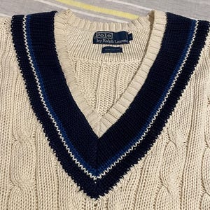 May include: Cream-colored cable knit sweater with a deep V-neck and navy blue and light blue striped trim. The sweater features the "Polo by Ralph Lauren" label. The knitwear has a classic, preppy style.