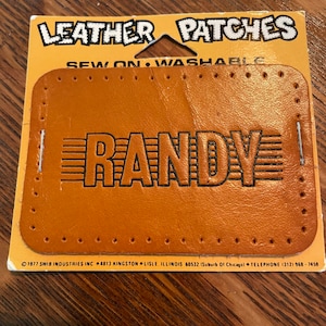 May include: A brown leather patch with the name "RANDY" in black lettering. The patch is sewn on and washable. The patch is from SWIB Industries Inc. and has a copyright date of 1977.