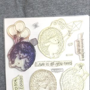May include: Clear acrylic stamps featuring hedgehog designs. One hedgehog holds balloons, another flies a kite, and others have various poses. Text includes "Love is all you need" and "Happy Now, Happy Forever."