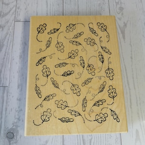 May include: A rectangular wooden stamp featuring a repeating pattern of stylized oak leaves and dotted lines. The leaves are black and arranged in a whimsical, flowing design across the light-colored wood surface. Ideal for crafting and scrapbooking.