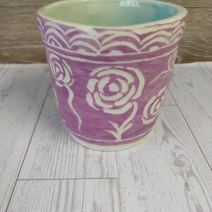 May include: A ceramic flower pot with a lavender exterior and a light green interior. The pot features white rose designs and a decorative border near the rim. The pot is sitting on a light-colored wooden surface.