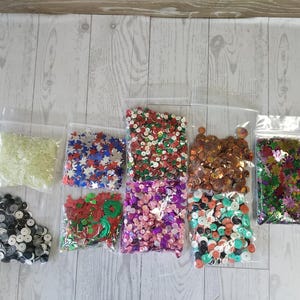 May include: A collection of various sequins in clear plastic bags. The sequins are in different shapes and colors, including stars, circles, and flower shapes. Colors include red, white, blue, green, gold, and iridescent.