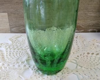 May include: A tall, green glass with a textured, bubbly design near the base. The glass is cylindrical and appears to be handmade. The glass is sitting on a white, crocheted doily.