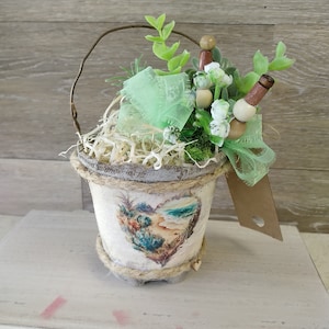 May include: A small white planter with a heart-shaped design featuring a beach scene. The planter is filled with green succulents, a green ribbon, and wooden beads. The planter has a wire handle.