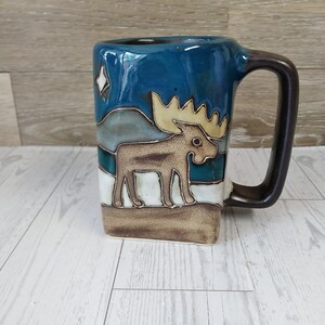 May include: A ceramic mug with a brown handle and a rectangular shape. The mug features a landscape design with a moose, mountains, and stars in shades of brown, blue, and white. The mug is approximately 4 inches tall.