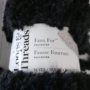 May include: A skein of black faux fur yarn with a white paper label. The label reads Loops & Threads Faux Fur Polyester Fausse Fourrure Polyester 56 YDS / 10.5 OZ / 51 M / 297 G. The yarn has a thick, fuzzy, and soft texture throughout the entire skein.