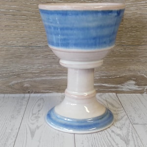 May include: A ceramic chalice with a flared blue and white bowl, a white stem, and a circular base. The bowl features horizontal blue stripes. The rim and base have a light pink accent.