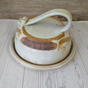 May include: A ceramic butter dish with a lid and handle. The dish is off-white with brown accents and the word "BUTTER" in brown lettering. The lid has a curved handle. The dish sits on a matching round base.