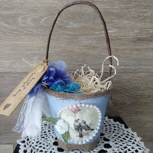 May include: A small blue and brown fabric basket with a brown paper handle. The basket is decorated with a floral design and two birds. The basket is filled with straw and a blue flower. The basket has a tag that says "Something old, something new".