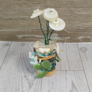 May include: A decorative object featuring a cylindrical base wrapped in blue, yellow, and white yarn, topped with a paper cutout. Three green wire stems extend upward, each ending in a white button flower. Small green leaves and white floral accents are attached to the side of the base.