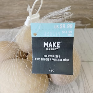 May include: A mesh bag containing seven DIY wood eggs. The eggs are a light beige color. A tag attached to the bag reads "MAKE MARKET DIY WOOD EGGS" and "ŒUFS EN BOIS À FAIRE SOI-MÊME".