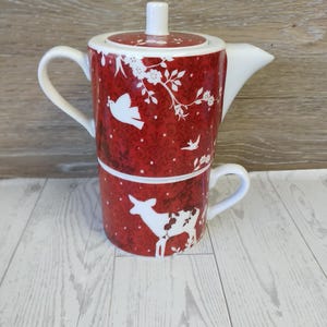 May include: A red ceramic teapot and teacup set with white floral and animal designs. The teapot has a lid and a spout. The teacup is stacked beneath the teapot. The set is on a light wood surface.