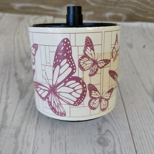 May include: A cylindrical container with a black top and a cream-colored body. The body is decorated with a pattern of purple butterflies and geometric shapes. The container sits on a light-colored wooden surface.