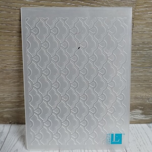 May include: A clear, rectangular embossing folder with a repeating, stylized scallop pattern. The folder has rounded corners and a Lifestyle Crafts logo in the lower right corner. The folder is made of a translucent plastic material.