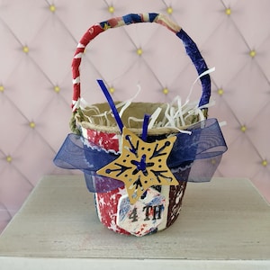 May include: A small, patriotic-themed basket with a handle wrapped in red, white, and blue fabric. The basket is decorated with a blue ribbon and a star-shaped ornament. The text "4 TH" is visible on the side of the basket.
