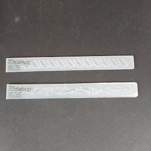 May include: Two clear, rectangular Cricut Cuttlebug embossing folders with a wave-like design. The folders have the Cricut Cuttlebug logo and text that reads "Organic Fragelah, Nineteen organic, Floritura Organica."