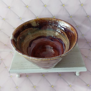 May include: A handmade ceramic bowl with a wide, open design. The bowl features a rich color palette of brown, burgundy, and tan, with a glossy finish. The bowl is displayed on a small, white, decorative stand.